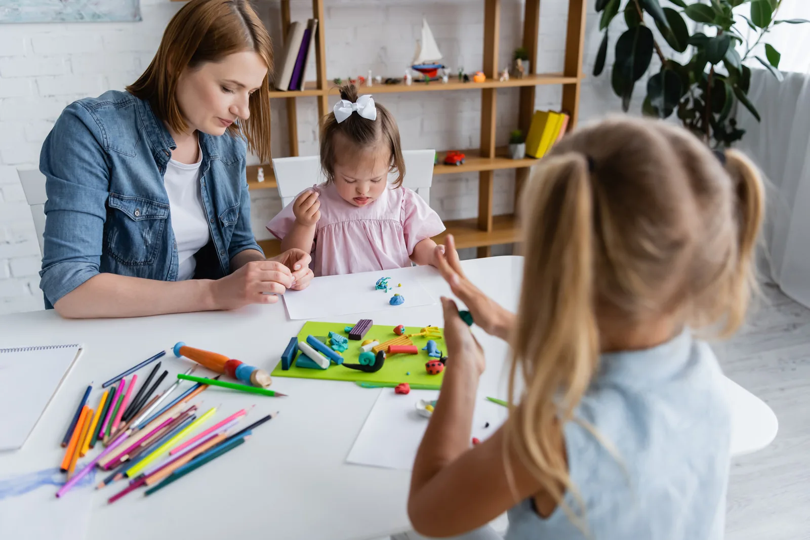 kindergarten teacher molding plasticine with disabled kid with down syndrome near blurred girl